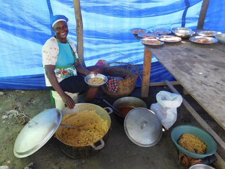 A smiling woman holds a vat of rice as she cooks on a stool with dishes around her on the ground. 