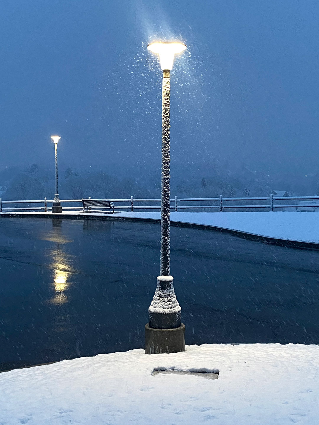 Two light poles, next to a glossy water-like parking lot, coated in fluffy snowflakes.