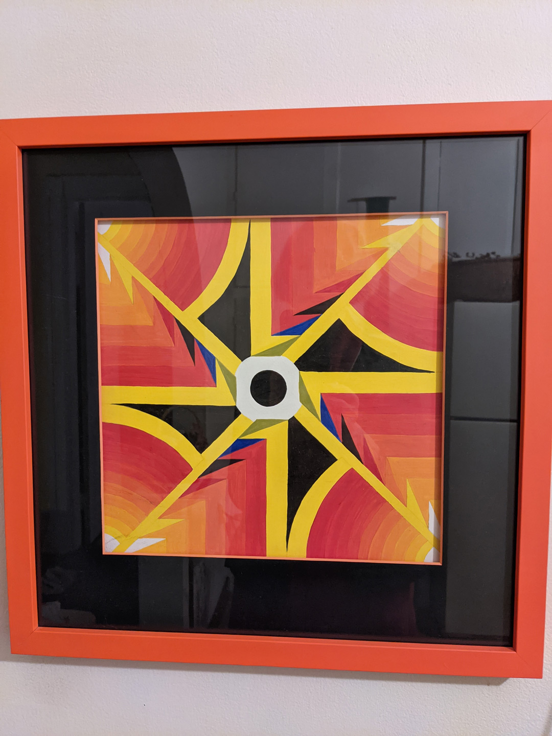 A graphic design of a spinning wheel in colors of orange, red, yellow, black and a little white and dark blue in a red frame with a black mat.