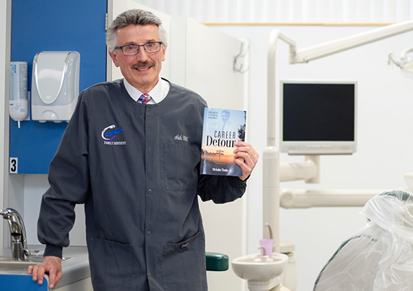 Nick stands in the college's dental hygiene lab, holding his book about his two career paths.