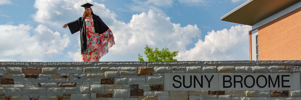 A young alumna in her graduation robe and cap standing on a stone wall that reads "SUNY Broome".